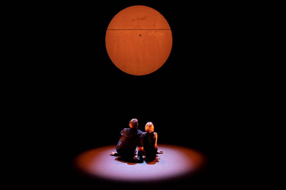 two dancers contemplate the golden orb as they sit huddled together in a small circle